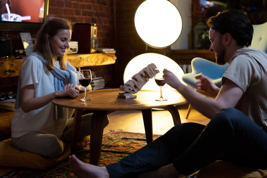 Smiling couple playing a stacking block game with glasses of red wine at a low table in a cozy, lamp-lit living room