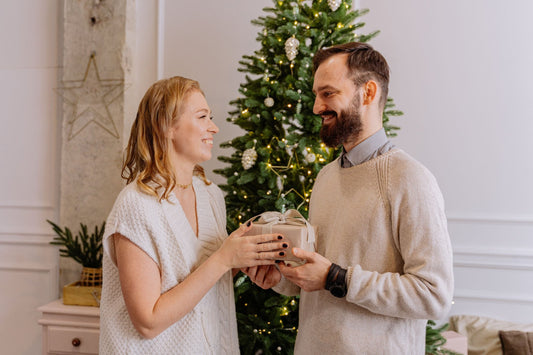 Smiling man handing his wife a small wrapped gift in front of a Christmas tree