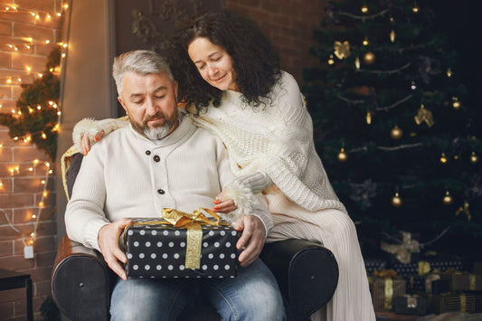 Man holding a wrapped gift while his wife hugs him beside the Christmas tree