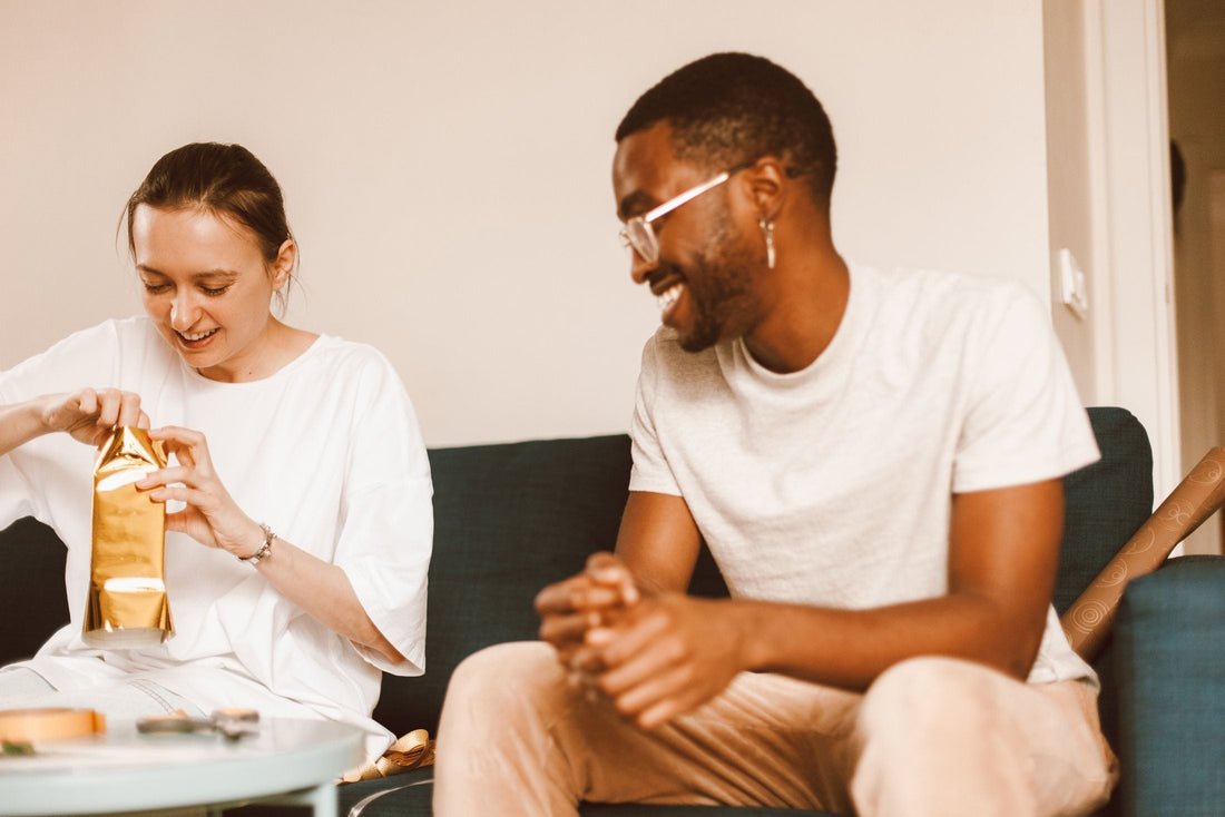Couple laughing on a sofa as she unwraps a gift in gold paper